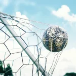 Sebuah bola sepak dengan desain grafis hitam putih tersangkut di jaring gawang sepak bola di bawah langit biru berawan.