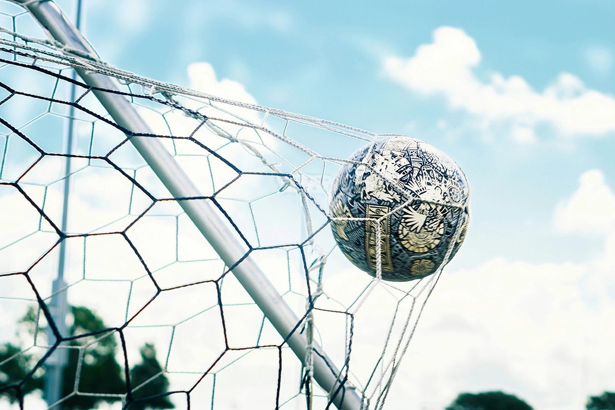 Sebuah bola sepak dengan desain grafis hitam putih tersangkut di jaring gawang sepak bola di bawah langit biru berawan.