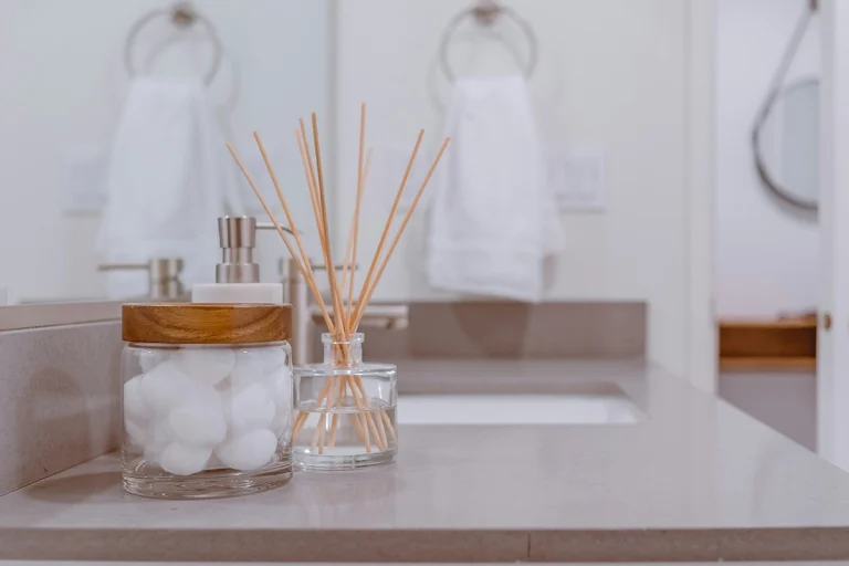 Foto close-up meja wastafel kamar mandi minimalis dengan reed diffuser berstik kayu dalam botol kaca dan wadah kaca berisi kapas. Latar belakang buram menunjukkan keran, cermin bundar, dan handuk putih yang digantung.