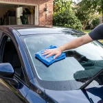 Seorang pria sedang mengelap kaca depan mobil sedan berwarna biru tua menggunakan kain microfiber biru dan cairan pembersih kaca, di depan garasi rumah.